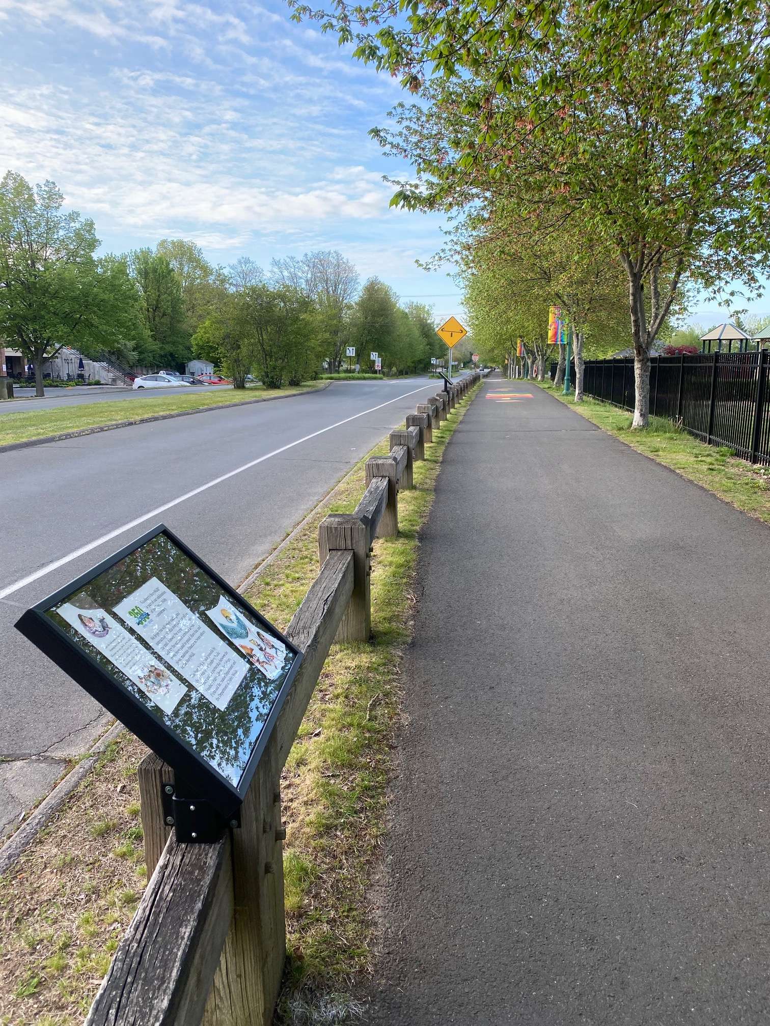 A StoryWalk frame rail-mounted to a wooden guardrail beside a sidewalk